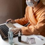 Woman with face mask working in cozy warm bath robe from home office surrounded by sanitizer, nasal drops,pills, thermometer. Ready for remote work on quarantine. Sick leave on remote online job