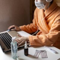 Woman with face mask working in cozy warm bath robe from home office surrounded by sanitizer, nasal drops,pills, thermometer. Ready for remote work on quarantine. Sick leave on remote online job Woman with face mask working in cozy warm bath robe from home office surrounded by sanitizer, nasal drops,pills, thermometer. Ready for remote work on quarantine. Sick leave on remote online job