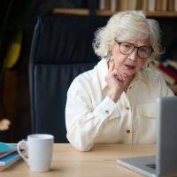 Attentiveness. Mature gray-haired elegant woman in glasses concentrated looking at laptop touching chin sitting at table indoors Attentiveness. Mature gray-haired elegant woman in glasses concentrated looking at laptop touching chin sitting at table indoors