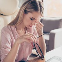 Pretty, charming, attractive woman tired from every day routine, holding fingers on nose between eyes, took off glasses, looking exhausted, sitting on armchair at desktop in work station