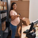 Young latin woman pregnant business worker scanning package label at office