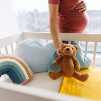 A pregnant woman in a pink dress places a teddy bear in a crib with a colorful rainbow pillow and blue cloud cushions, creating a warm, nurturing atmosphere. A pregnant woman in a pink dress places a teddy bear in a crib with a colorful rainbow pillow and blue cloud cushions, creating a warm, nurturing atmosphere.