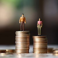 Miniature businessman standing on a taller stack of coins than a businesswoman, illustrating the gender pay gap.